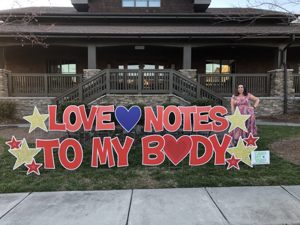 Nicole C. Ayers stands beside a colorful sign that says Love Notes to My Body.